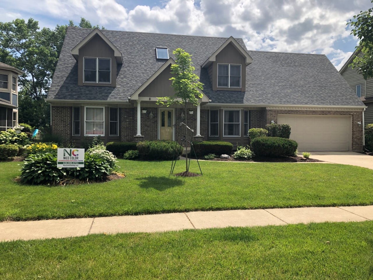 A suburban two-story home with dark brown siding, dormer windows, and a Nice Color Painting yard sign on the front lawn