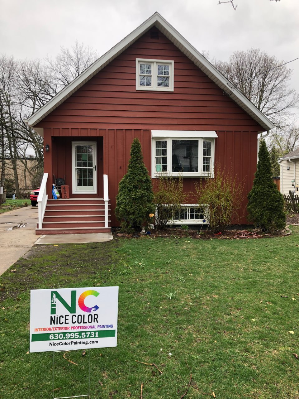 A two-story house with deep red siding, white trim, and a Nice Color yard sign in the front lawn